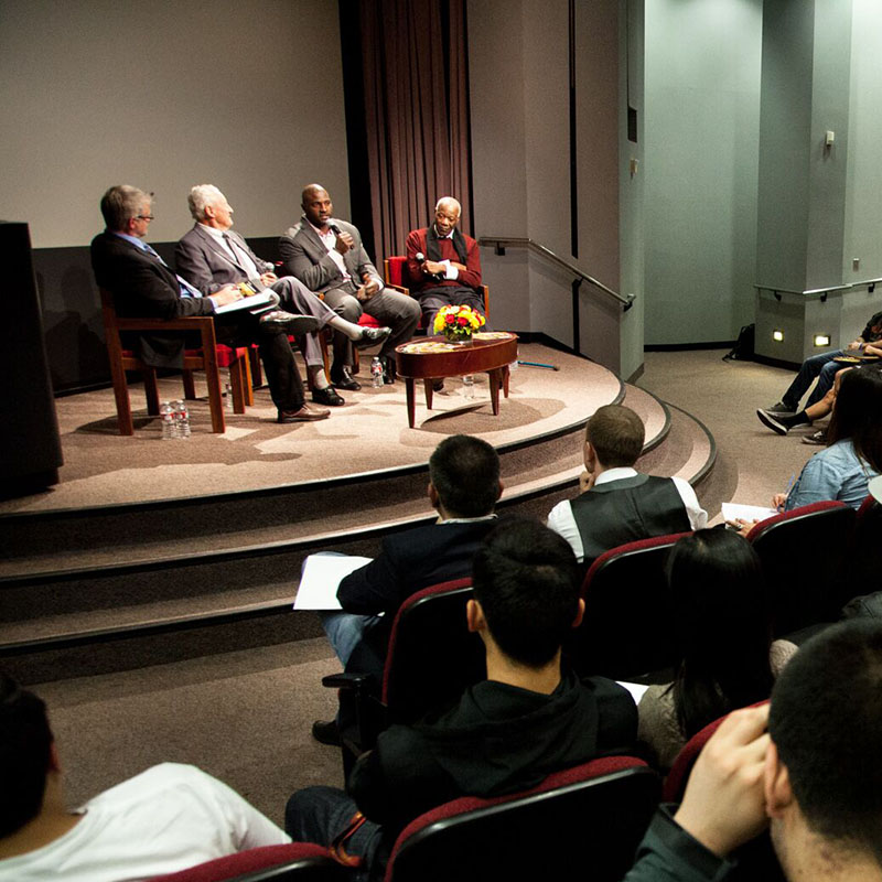 Communication students attending a panel discussion in an auditorium with four panelists on the stage and one speaking on a wireless microphone